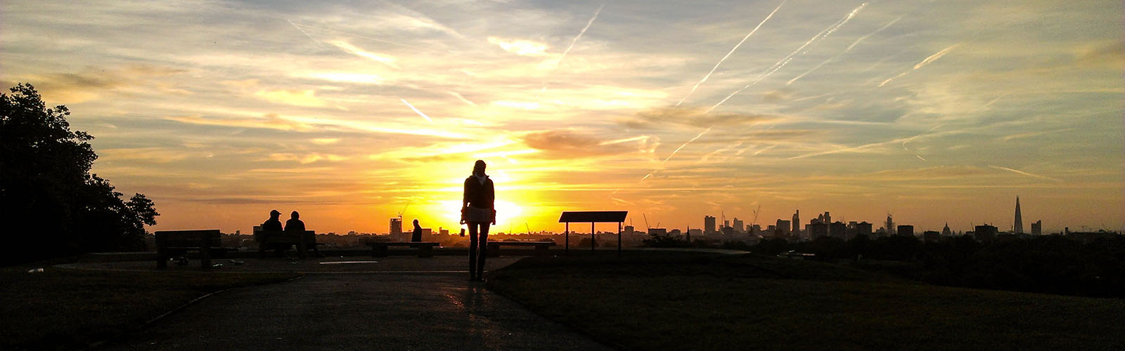 A student is facing towars a sunset on Primrose Hill. The London City skyline is in the background and covered by a shadow from the sun.